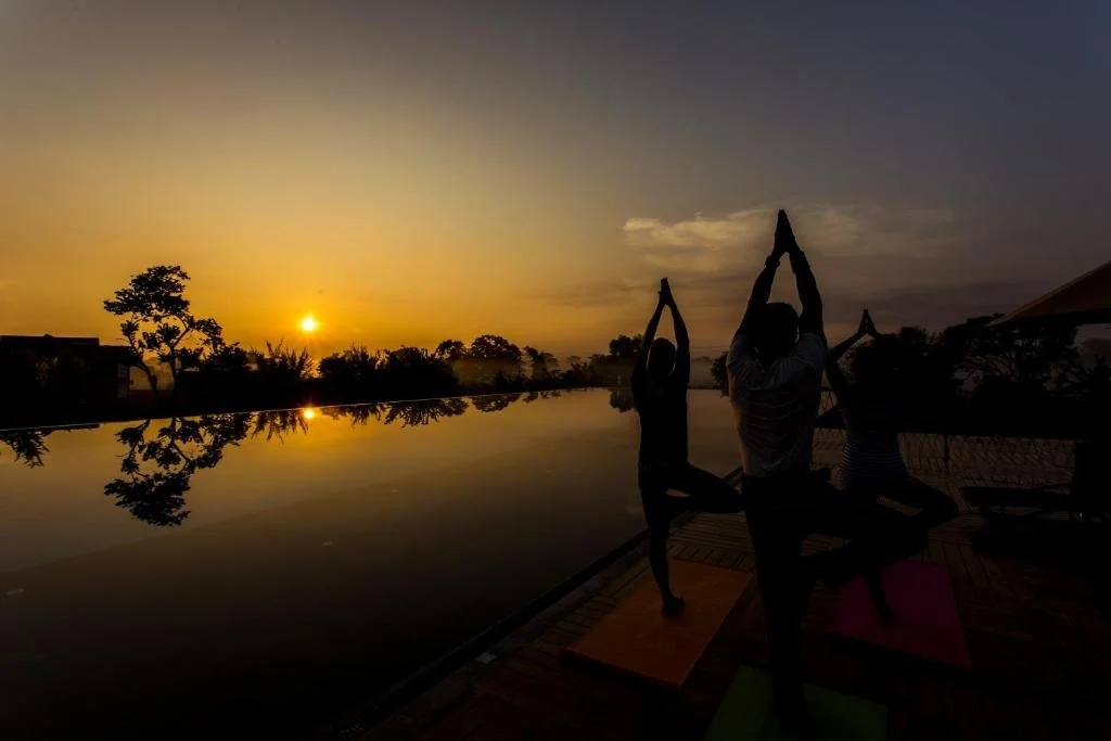 Silhouettes of three people practicing yoga at sunset near a calm body of water with trees and buildings in the background.