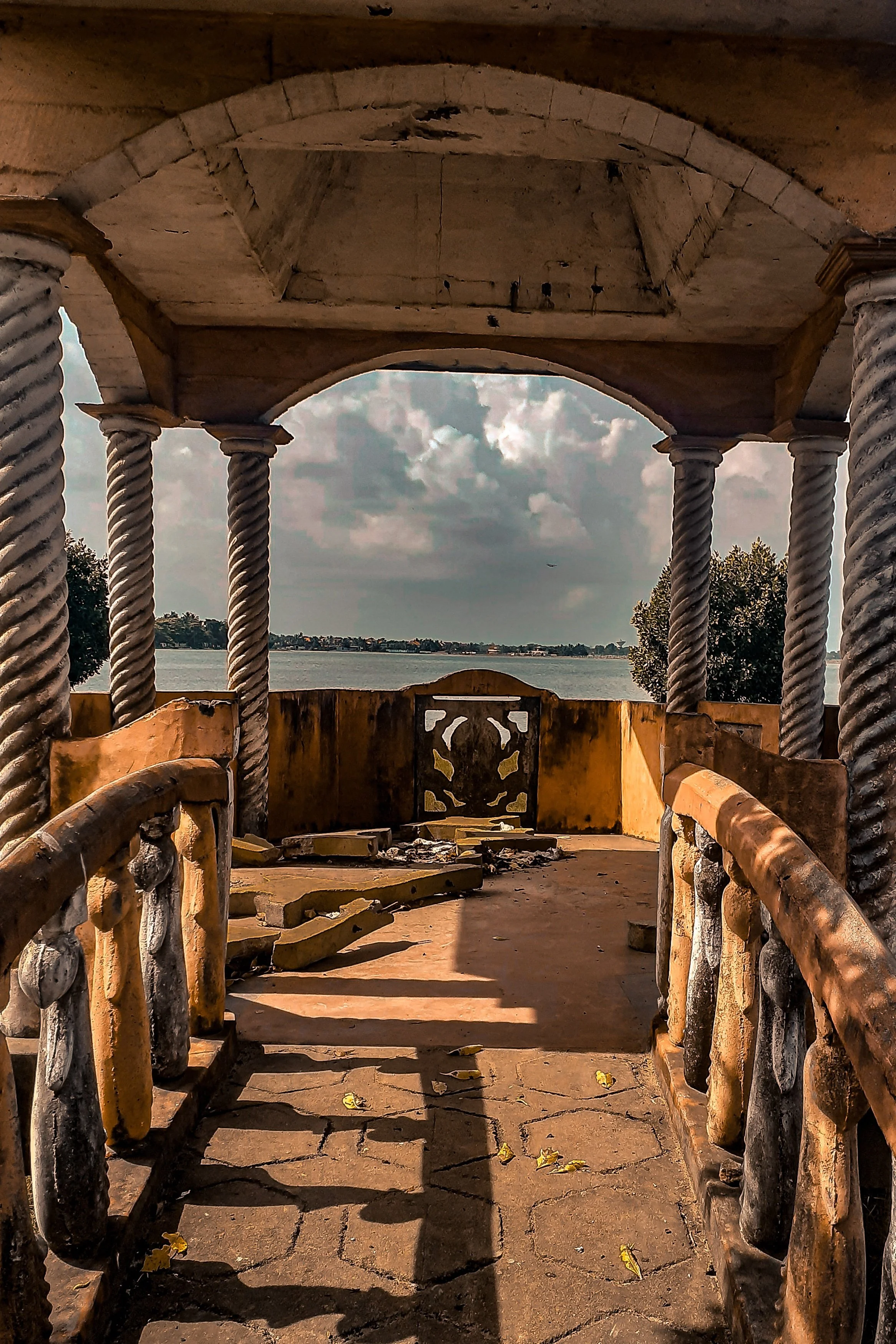 View through the stone and wood railing of an abandoned, decorative balcony or gazebo overlooking a body of water with trees and a cloudy sky in the distance, in Sri Lanka