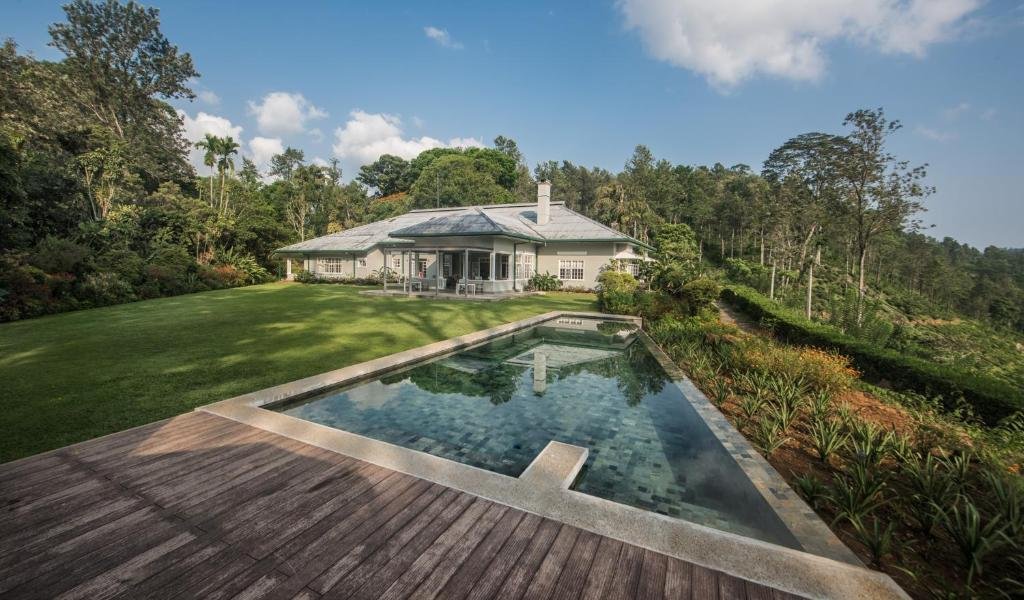 Large house with a light gray exterior, surrounded by lush green grass and trees, with a rectangular swimming pool in the foreground and a cloudy sky overhead.