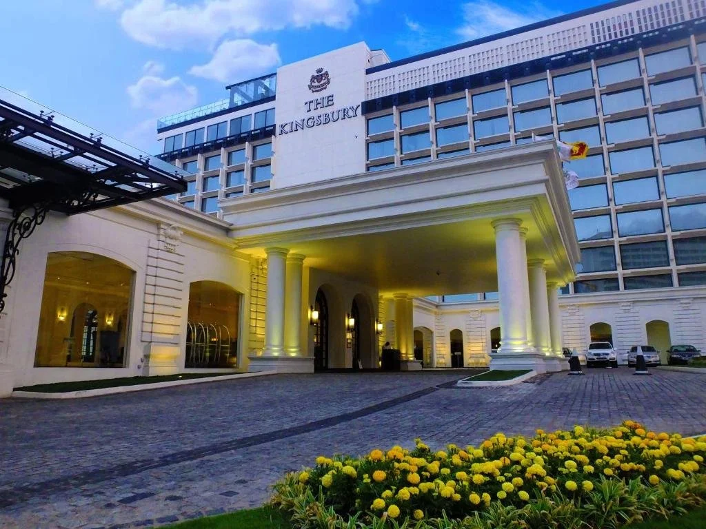 The exterior of The Kingsbury hotel with a parking area and yellow flower bed in the foreground during dusk.