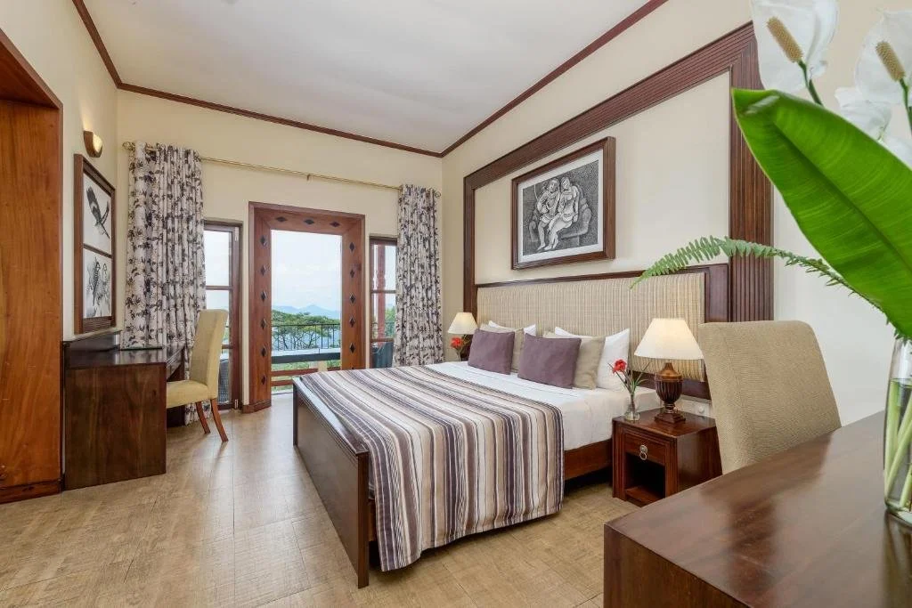 Hotel room with a large bed, nightstands, a desk with a chair, and a balcony with a view of mountains and greenery.