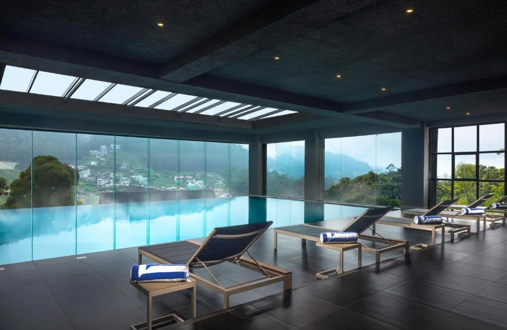 Indoor swimming pool with large windows showing a hilly landscape, surrounded by lounge chairs and rolled towels.