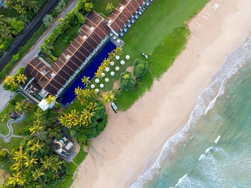 Aerial view of a beachfront resort with red-roofed buildings, a swimming pool, palm trees, and white umbrellas on the beach.