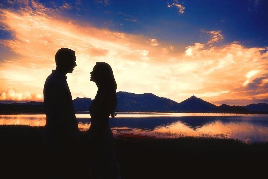 Silhouetted couple facing each other near a lake at sunset, with mountains and colorful sky in the background.