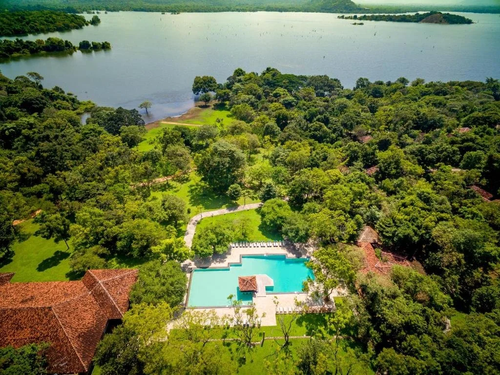 Aerial view of a luxury resort with a large swimming pool surrounded by trees and grass, near a river or lake with small islands.