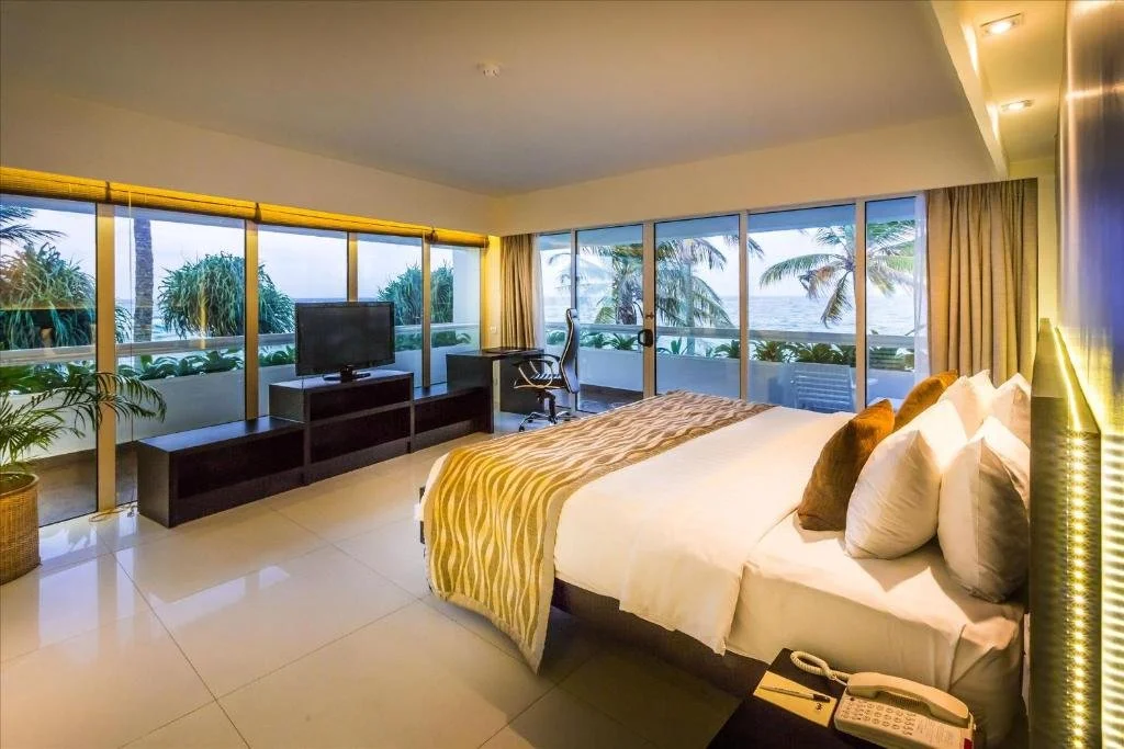 Modern hotel room with a large bed, pillows, and a balcony view of palm trees and the ocean.