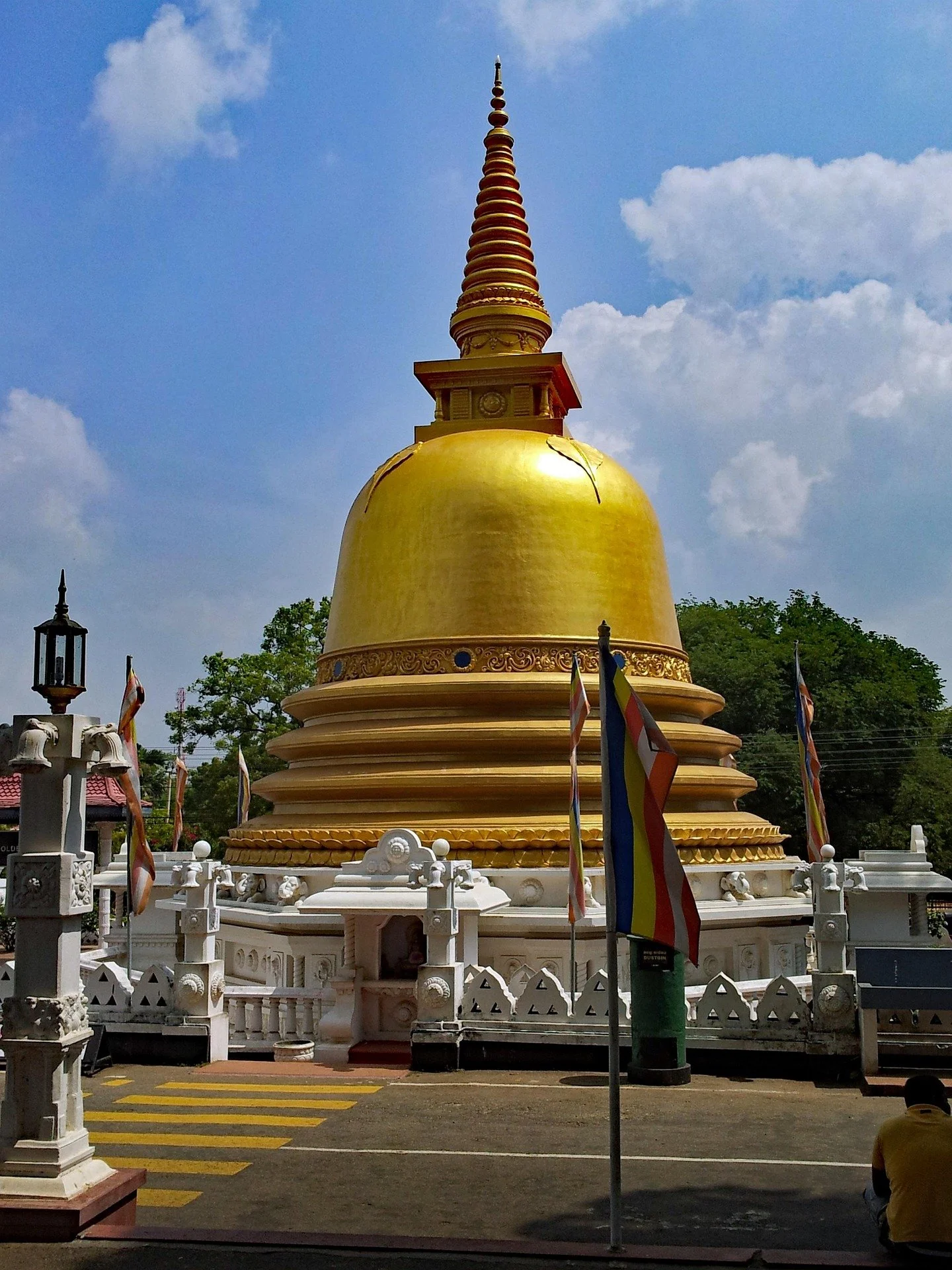 A large golden Buddhist stupa with a tall, multi-tiered spire, surrounded by flags and a white fence, against a cloudy blue sky.