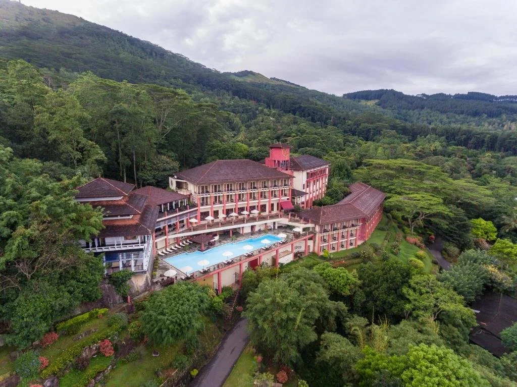 A large hotel with multiple levels and balconies, surrounded by lush green trees and mountainous terrain, with a swimming pool in front.
