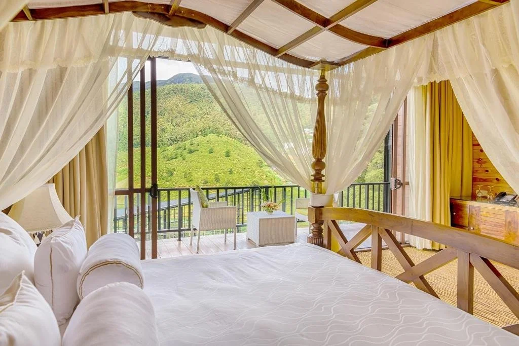 A bedroom with a four-poster bed, white bedding, and cream-colored curtains, opening to a balcony with a mountain view.