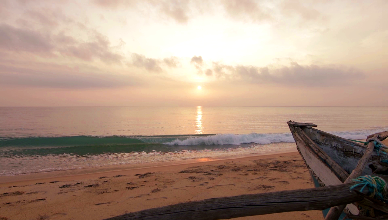 Sunset over calm ocean with small waves hitting sandy beach and weathered wooden boat on shore