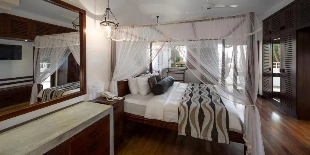 A bedroom with a four-poster bed draped with white mosquito netting, wooden furniture, a large mirror, a bedside table with a phone, and a window with white curtains.