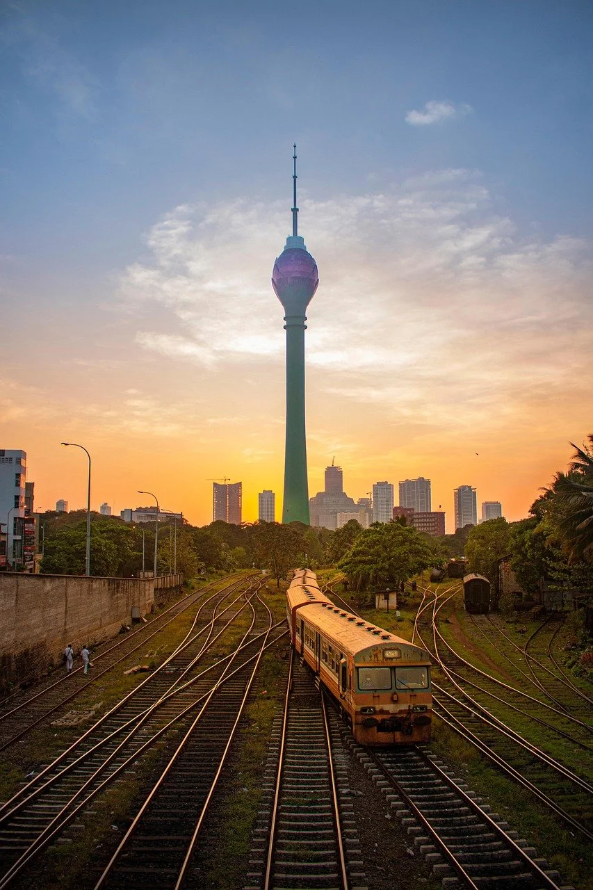 A railway train on multiple tracks heading towards a city skyline during sunset, with a tall television tower in the background.