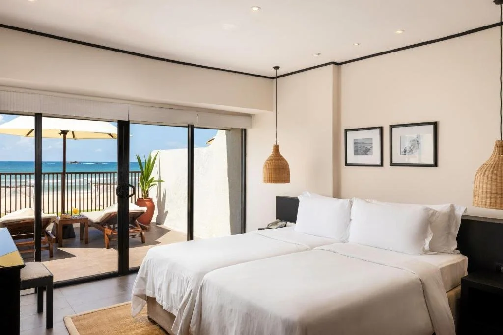 A hotel room with a large bed, hanging wicker lamps, and framed black-and-white photos on the wall. The room opens to a balcony with lounge chairs, a table, and an umbrella, overlooking a beach and ocean.