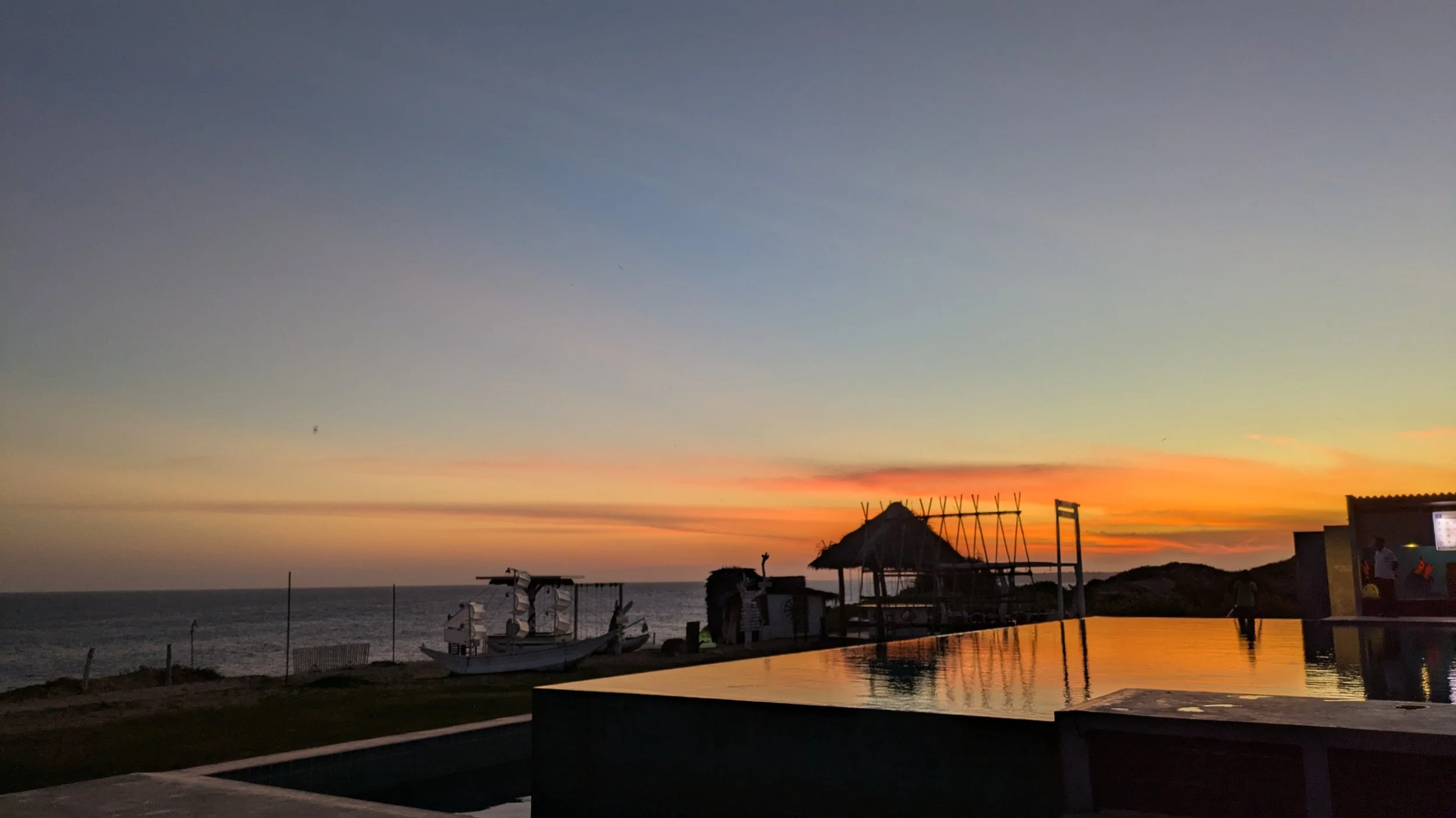 Sunset over the ocean with silhouettes of boats and structures, and a reflection of the colorful sky on a pool's surface.