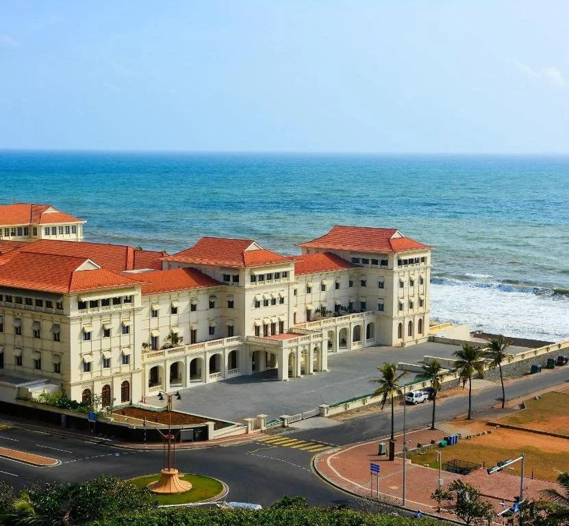 A large luxury hotel with red-tiled roofs on a beachfront property, overlooking the ocean with waves crashing on the shore, surrounded by palm trees and a paved road.