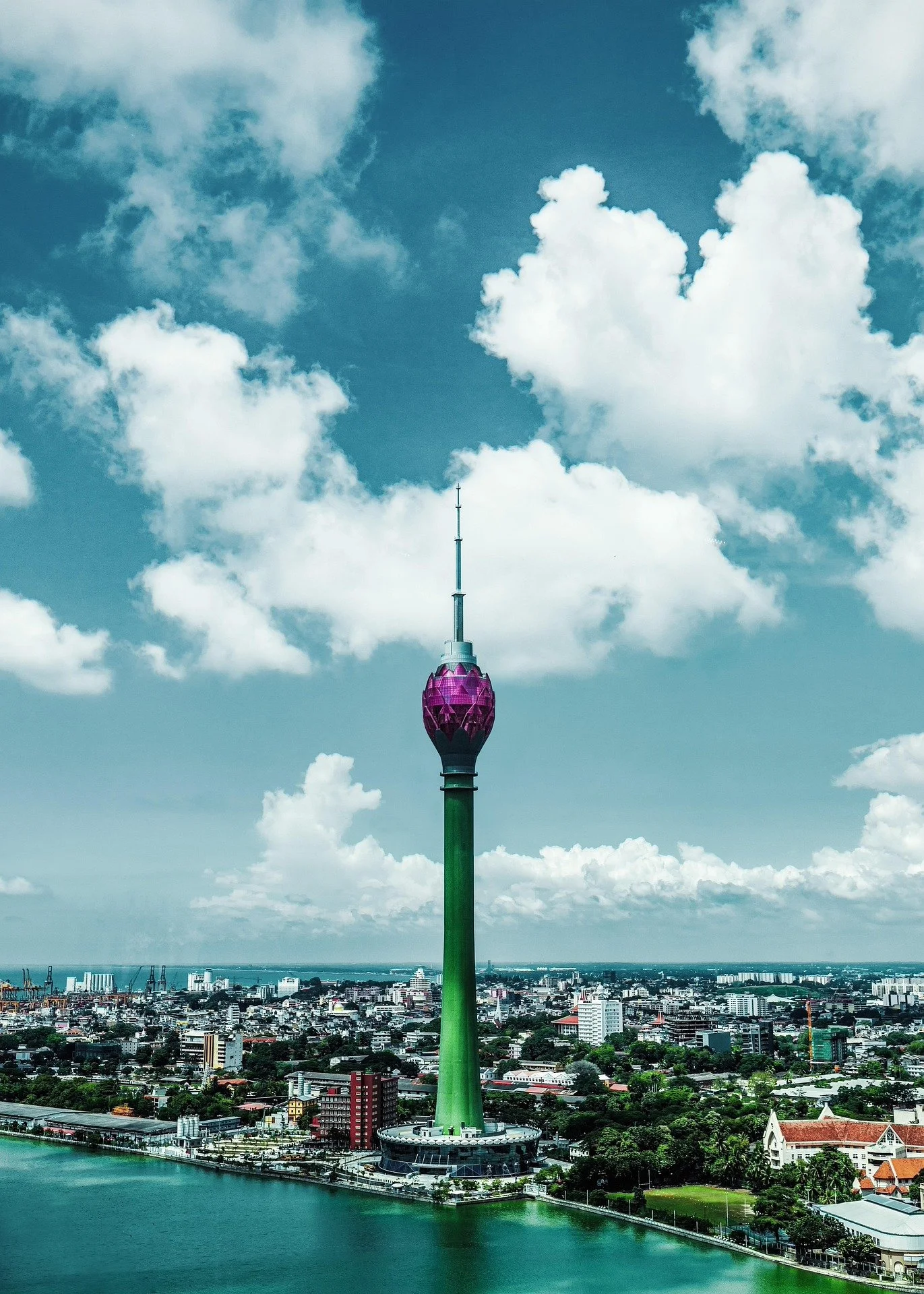 A tall green tower with a purple top stands over a city with water in the foreground and partly cloudy sky.