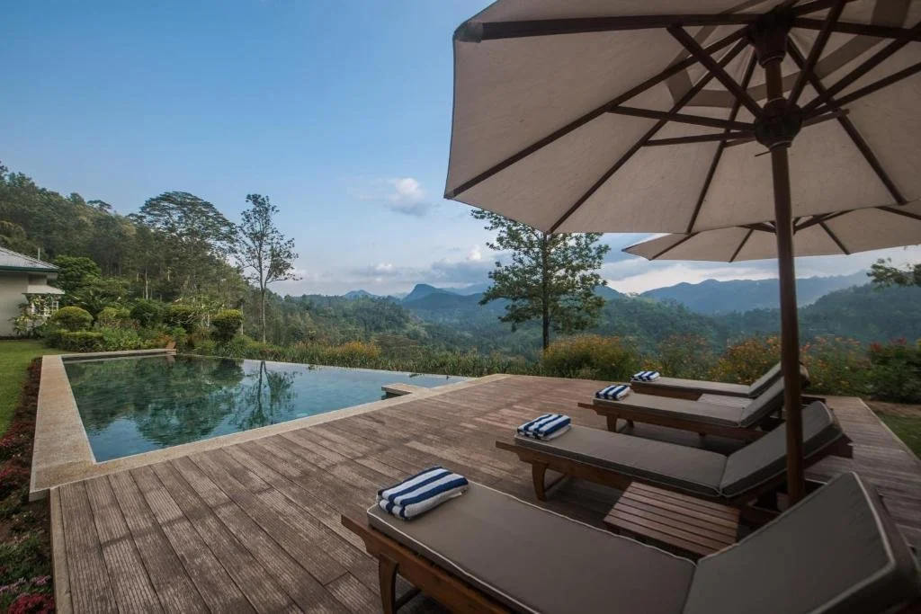Outdoor pool area with lounge chairs, an umbrella, and a mountain landscape in the background.