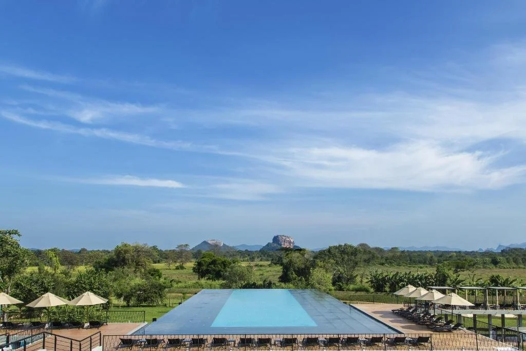 Outdoor swimming pool surrounded by lounge chairs and umbrellas, with lush green landscape and distant mountains under a blue sky with scattered clouds.