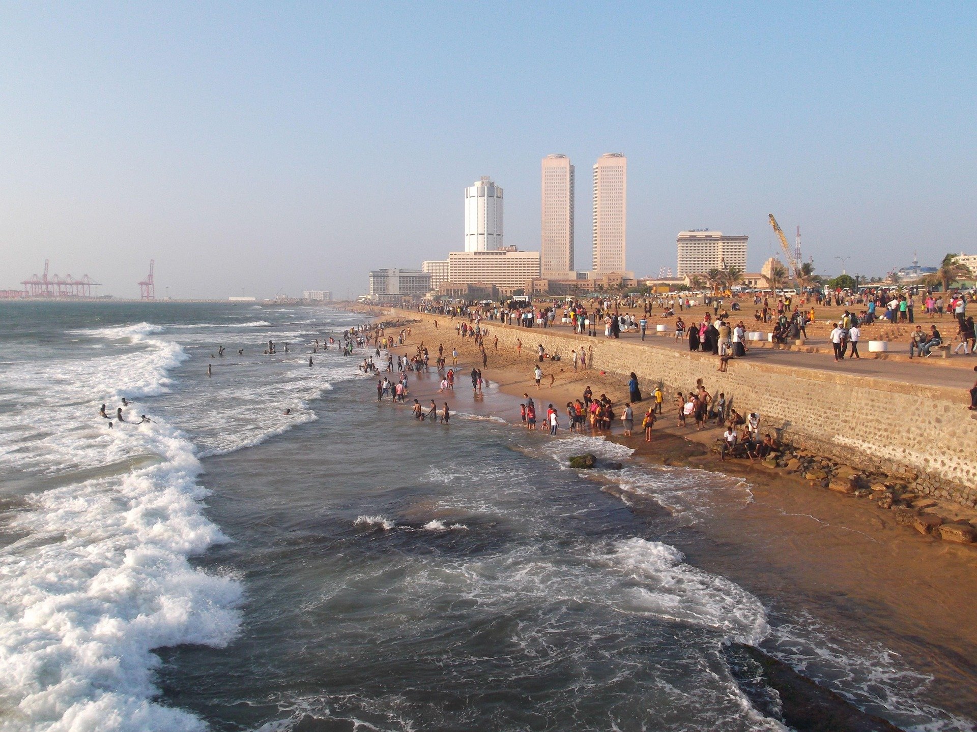 A crowded beach with people swimming and playing in the ocean, a promenade with people walking and sitting, high-rise buildings and construction cranes in the background.