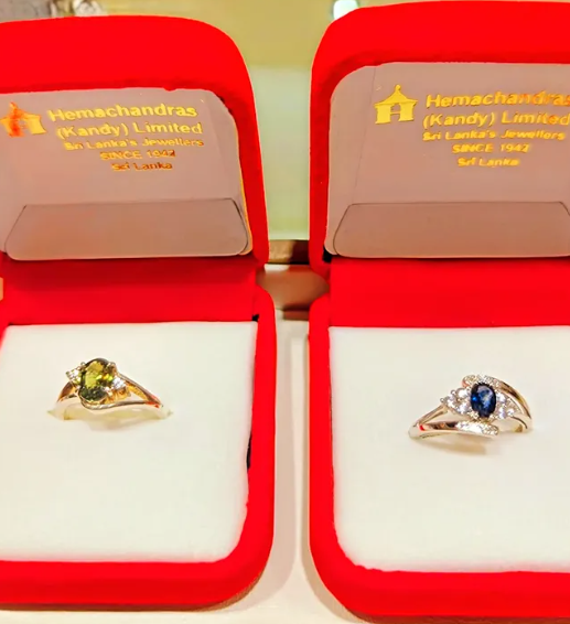 Two rings in red jewelry boxes, one with a yellow gemstone and the other with a dark blue gemstone, both surrounded by smaller clear stones.