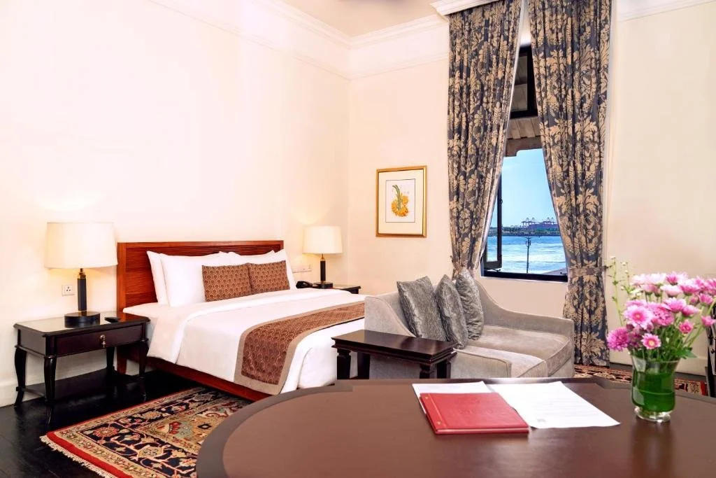 Hotel bedroom with a large bed, bedside tables with lamps, a sofa with cushions, a framed artwork on the wall, a window with blue patterned curtains, and a vase of pink flowers on a table.