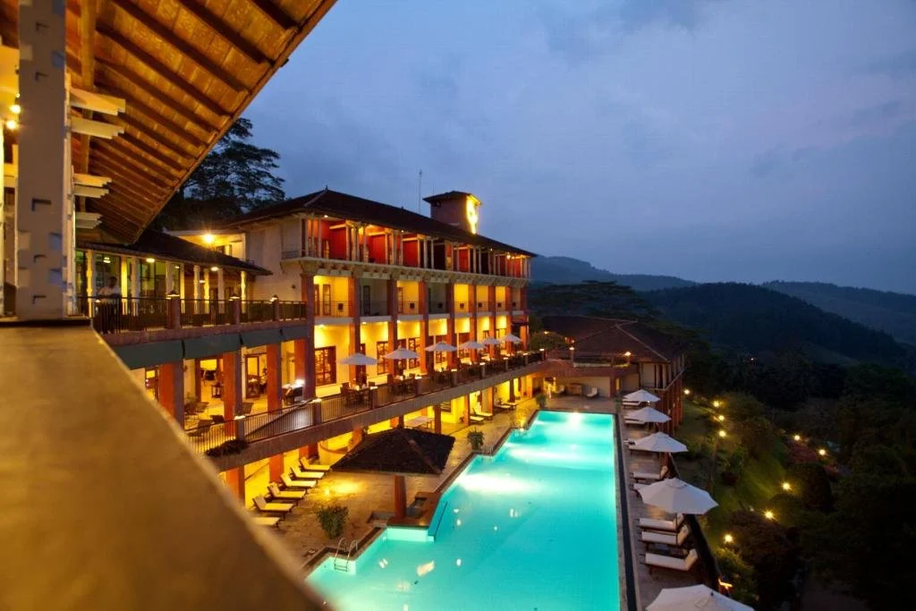 A multi-story hotel with outdoor swimming pool, illuminated during evening, overlooking a hilly landscape with trees and distant lights.