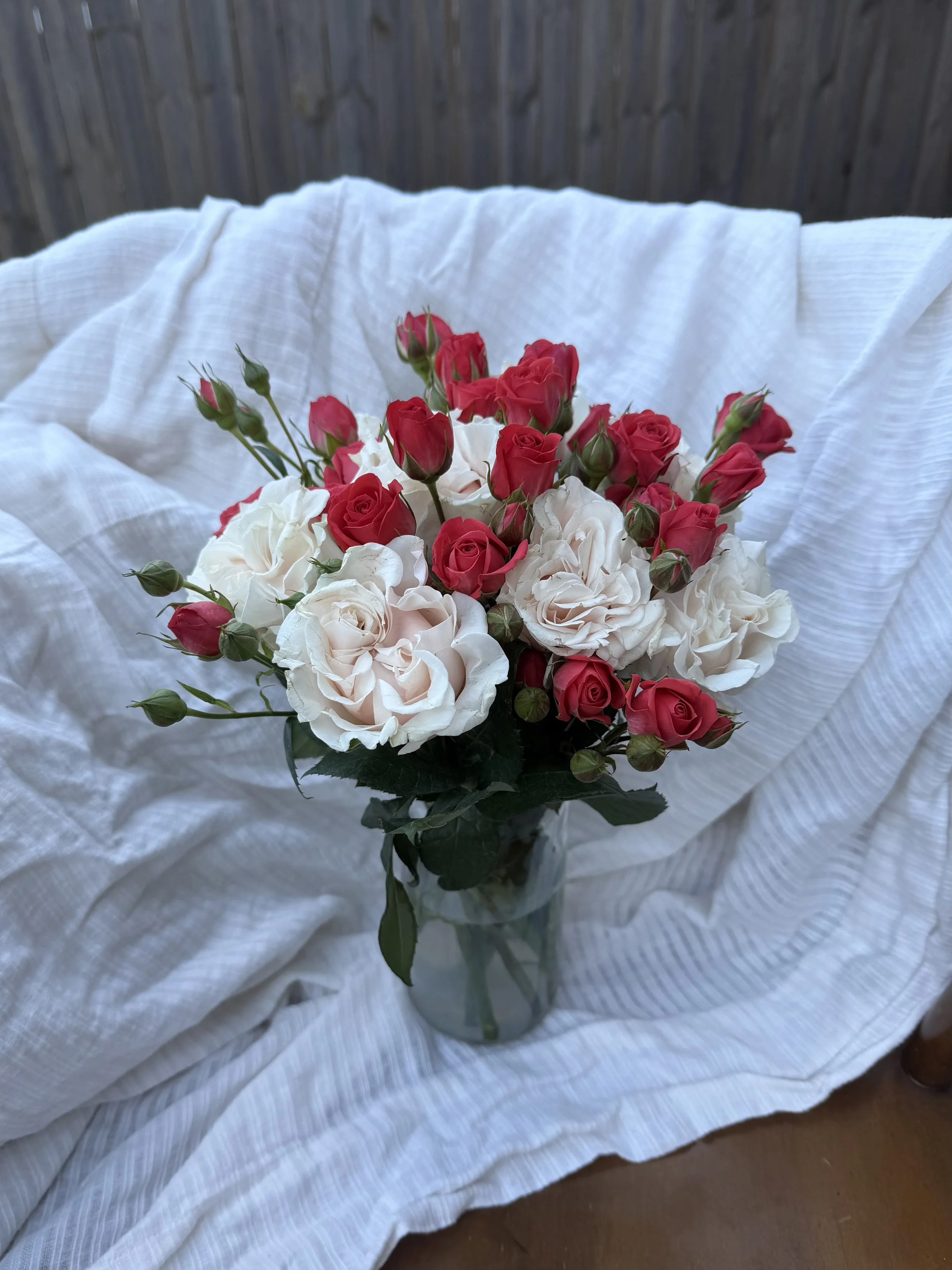 A bouquet of pink and white roses in a glass vase on a white cloth with a wooden fence background.