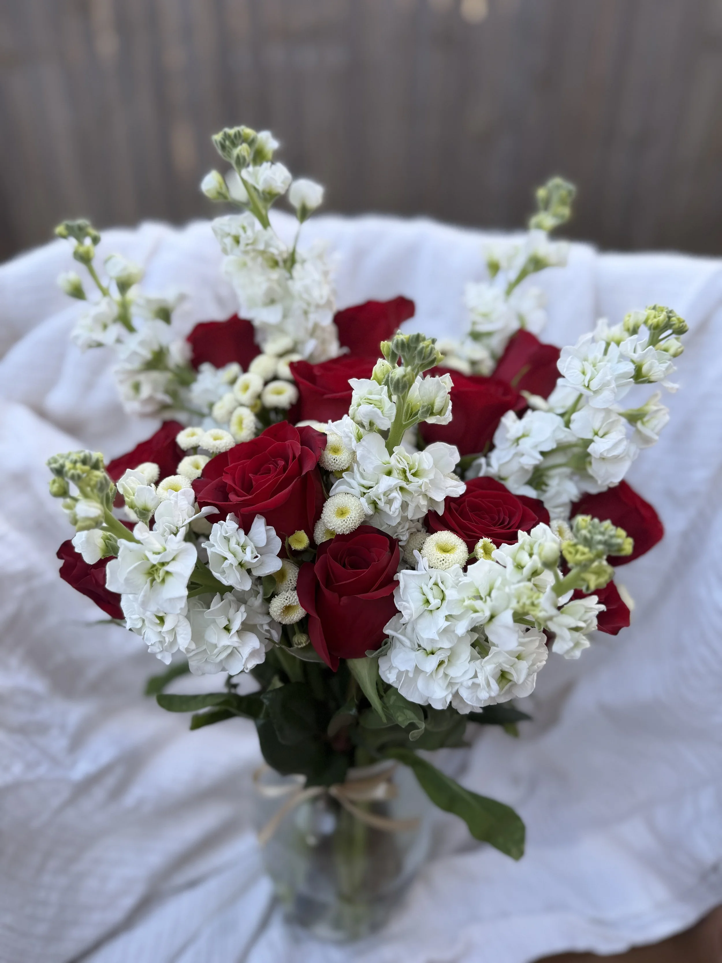 A bouquet of red roses, white stock, and white baby's breath flowers in a glass vase, resting on white fabric.