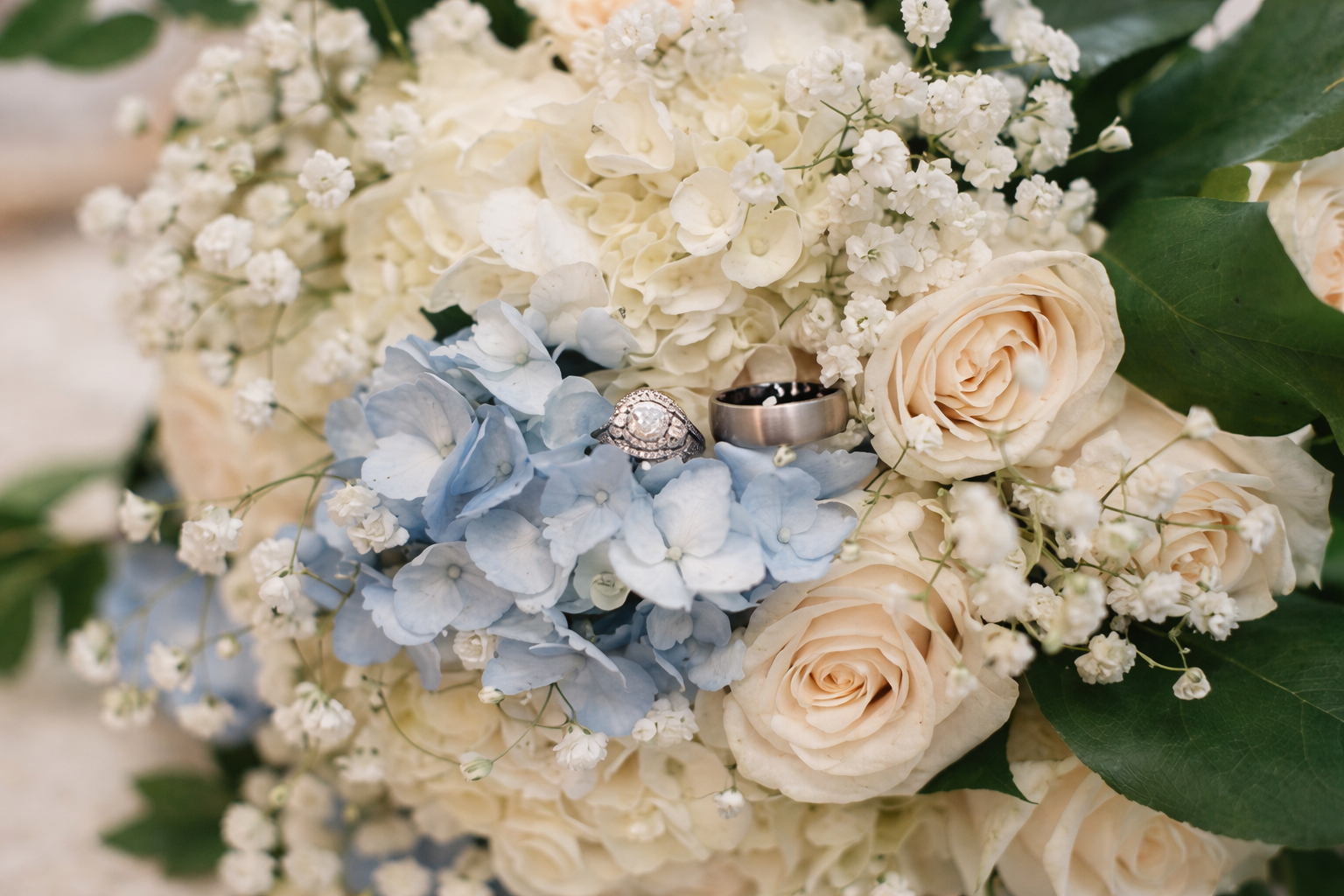 A wedding bouquet consisting of white roses, blue hydrangeas, baby's breath, and green leaves, with a diamond engagement ring and a silver wedding band placed among the flowers.
