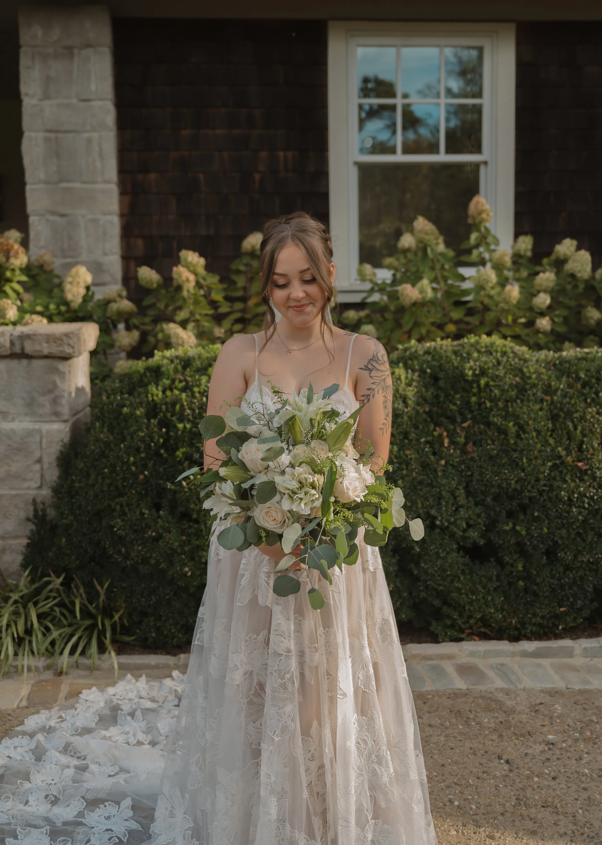 A bride in a lace wedding dress holding a bouquet of white and green flowers outdoors, smiling gently with a garden and house in the background.