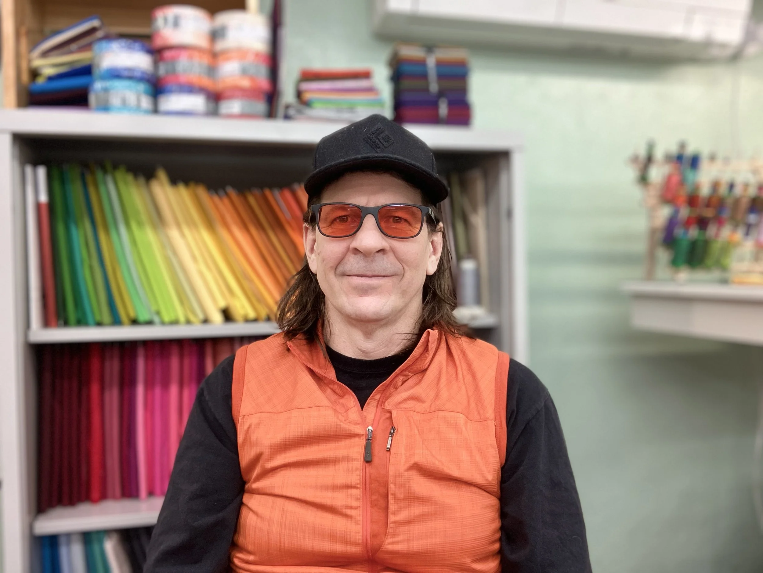 A man with long hair, wearing an orange vest, black cap, and red sunglasses, sitting in front of colorful shelves in a store.