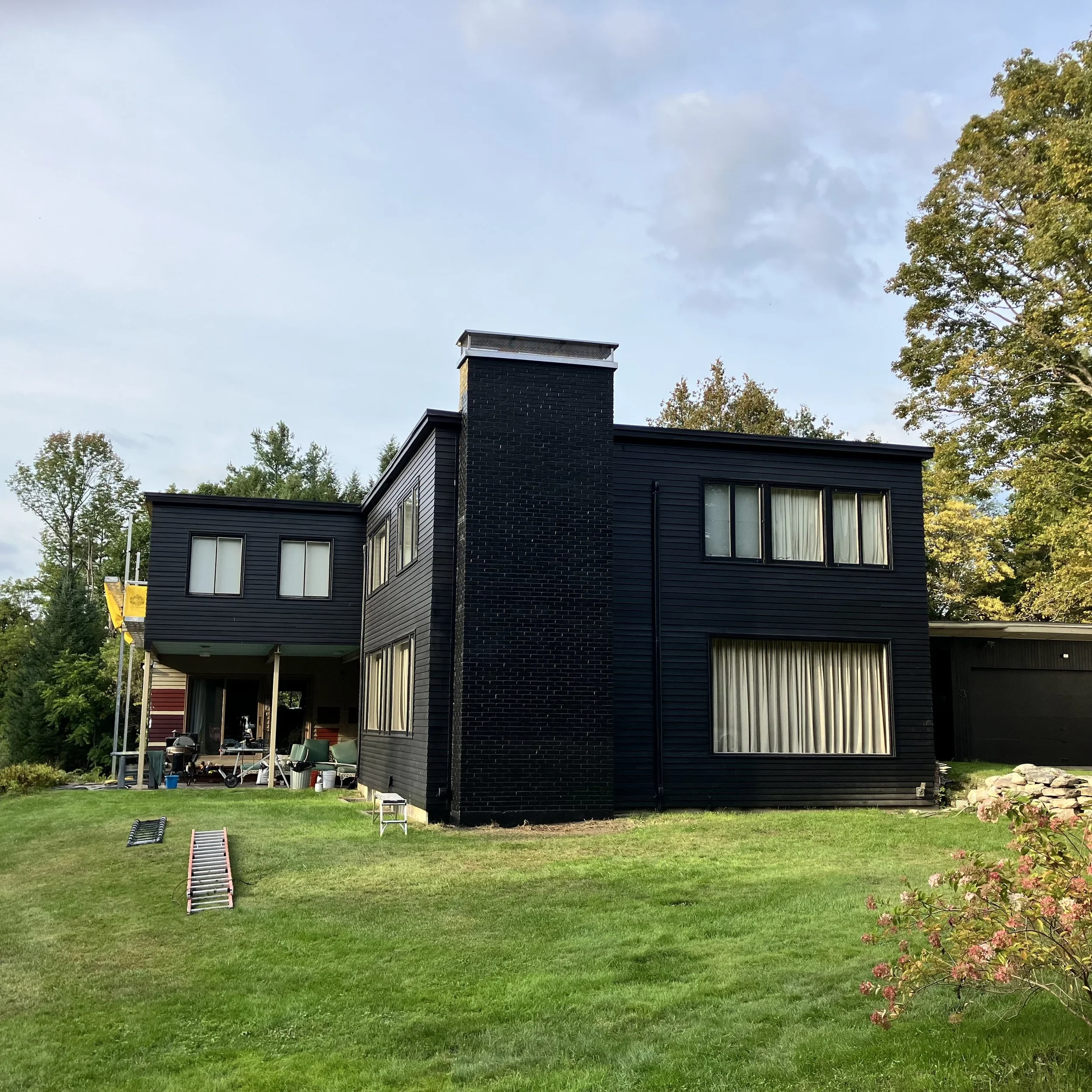 Black modern two-story house with large windows, surrounded by a green lawn and trees, under a partly cloudy sky.