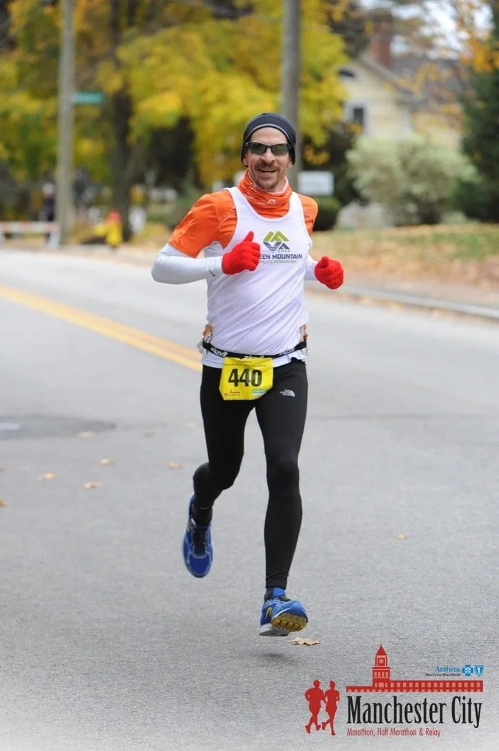 A man running outdoors during a marathon race, wearing sunglasses, a black headband, a white and orange athletic shirt, black running tights, blue running shoes, red gloves, and a yellow race bib number 440. The background has trees with fall foliage