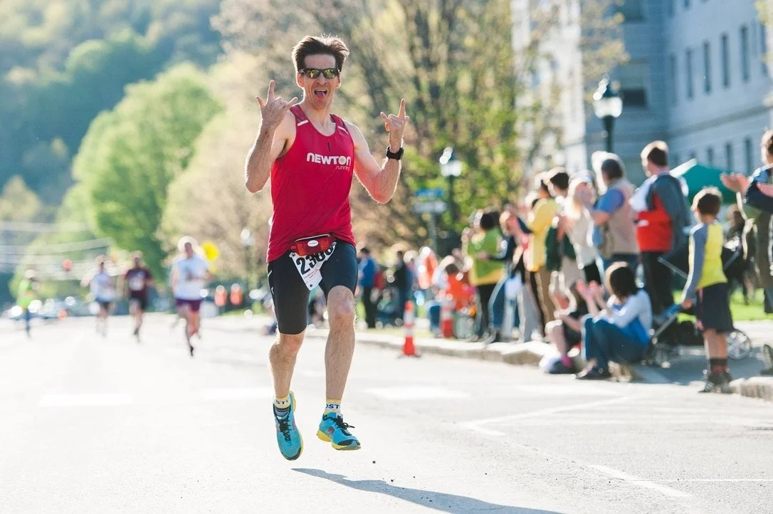 Male runner wearing sunglasses, a red tank top, and black shorts, crossing the finish line in a marathon with a crowd cheering on the sidelines.
