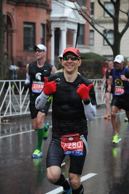 Marathon runner smiling and giving a thumbs-up during a race on a rainy day, with other runners and city buildings in the background.