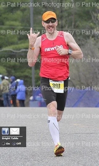 A male marathon runner wearing a red tank top, black shorts, yellow shoes, an orange cap, and sunglasses, running outdoors during a race, making a hand gesture and smiling.
