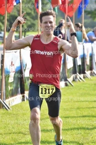 Male runner in a red tank top and black shorts crossing the finish line, holding his arms up in a victory pose during a race event.