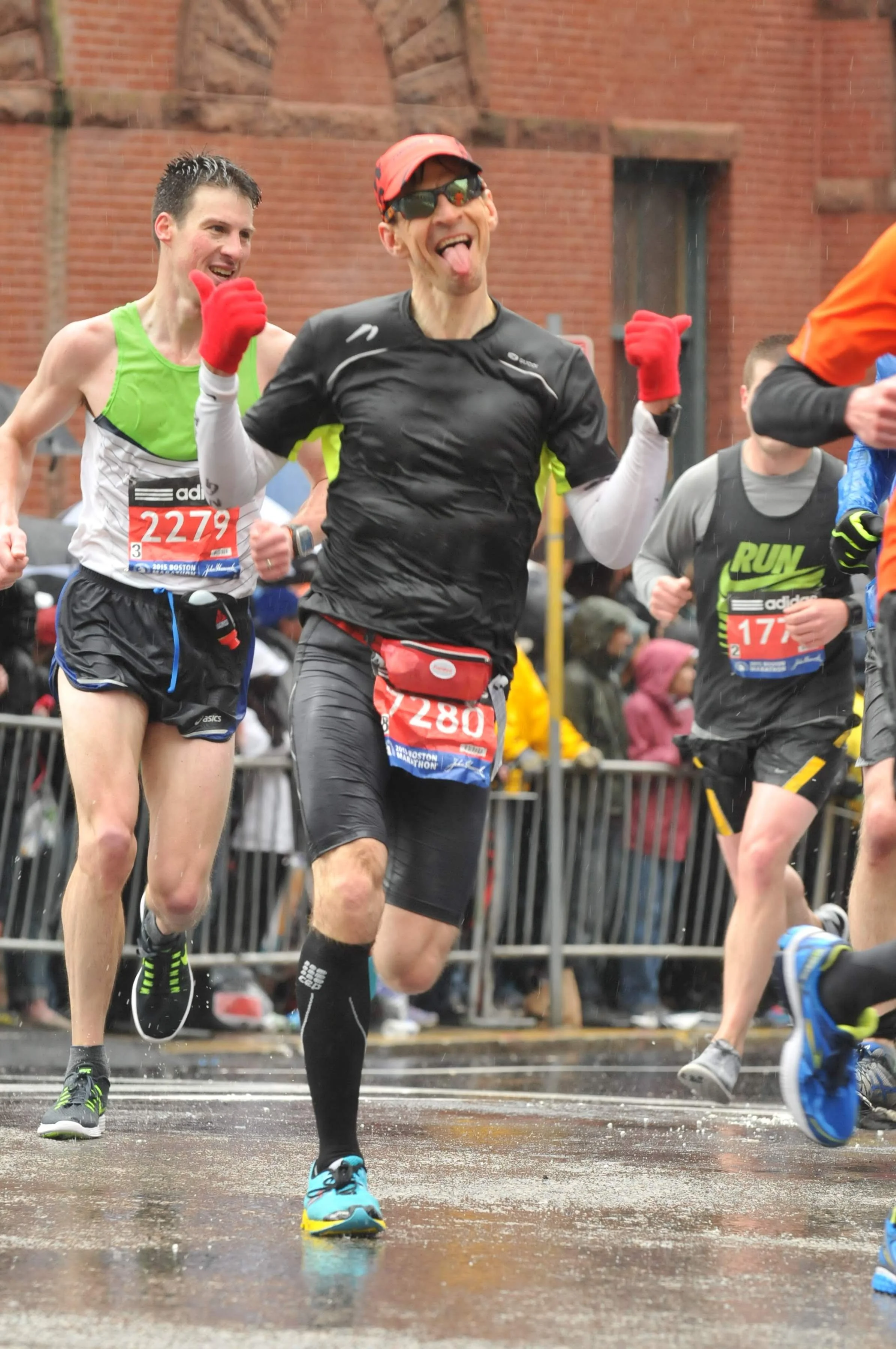Marathon runners finish line, smiling, with wet street and spectators behind barriers