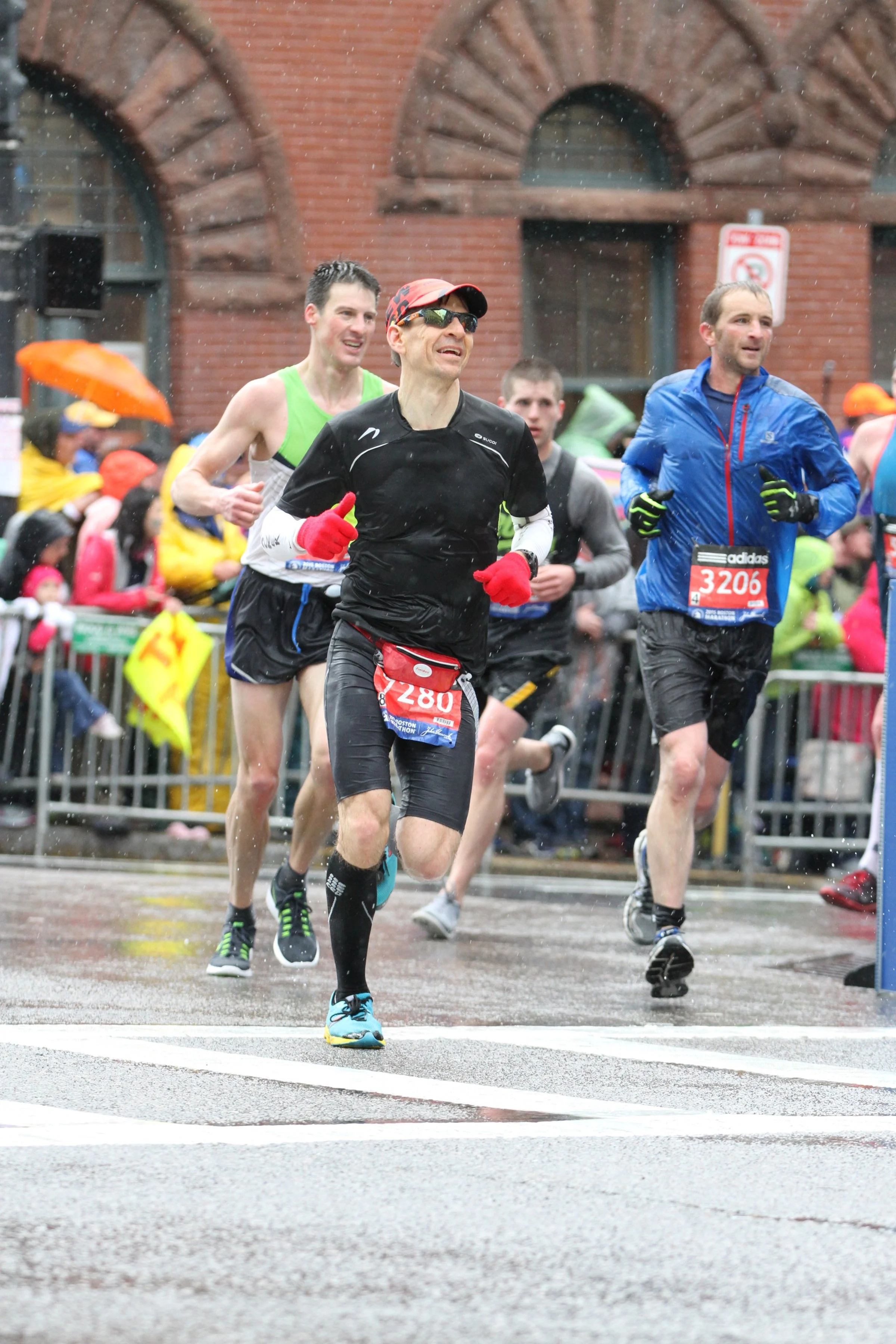 Group of runners participating in a marathon or race on a rainy day, with spectators behind barriers and a brick building in the background.
