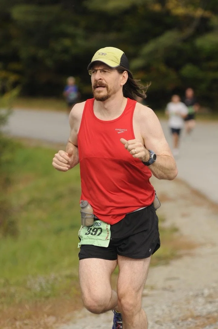 A man running outdoors on a trail, wearing a red sleeveless shirt, black shorts, a yellow and black cap, glasses, and a running watch. He has a race bib with the number 397 attached to his shorts and appears focused while running.