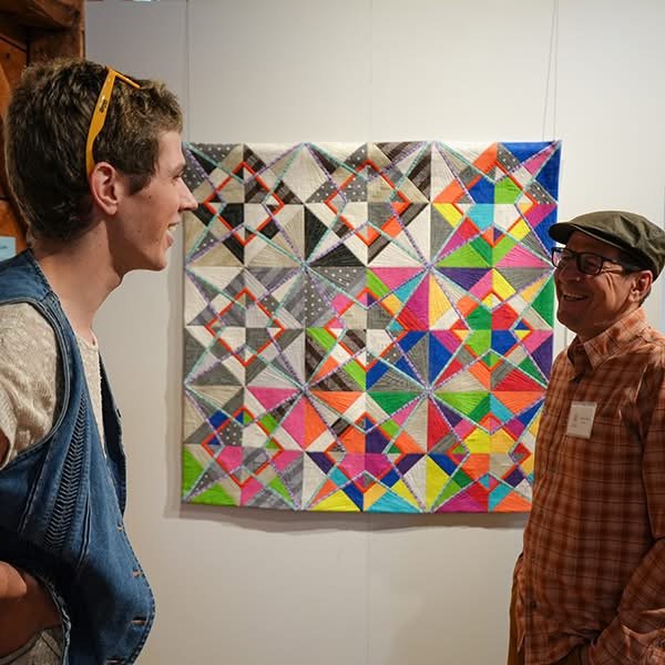 Two men smiling and talking in front of a colorful quilt displayed on a wall.