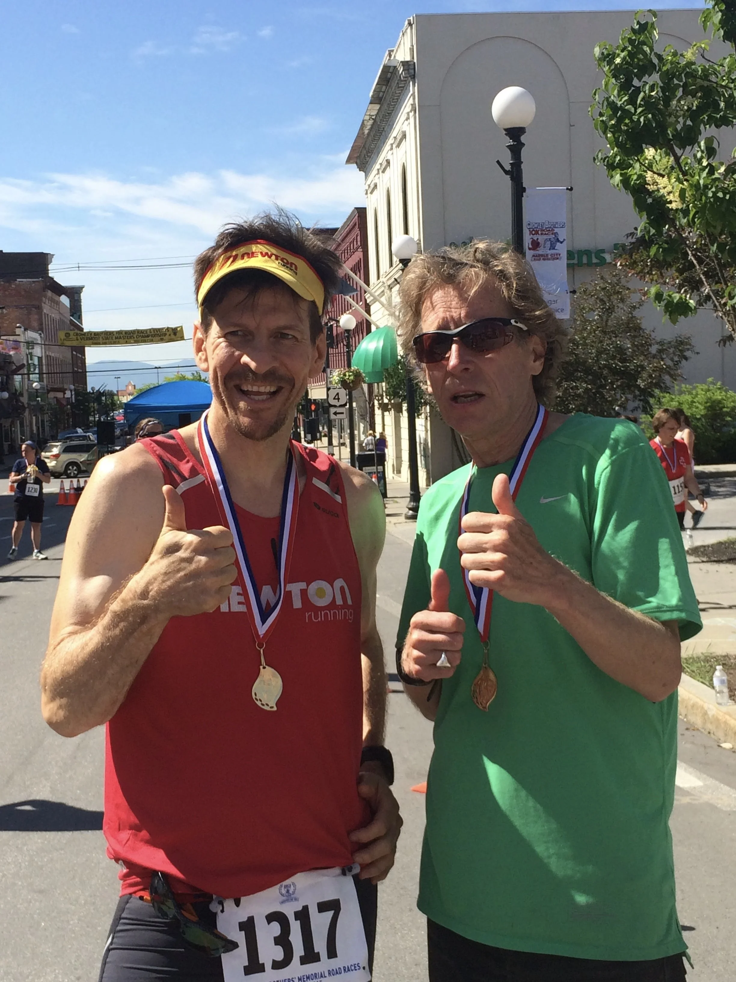 Two men showing medals after running a race, giving thumbs up, standing on a city street with other runners in the background, buildings, and trees.