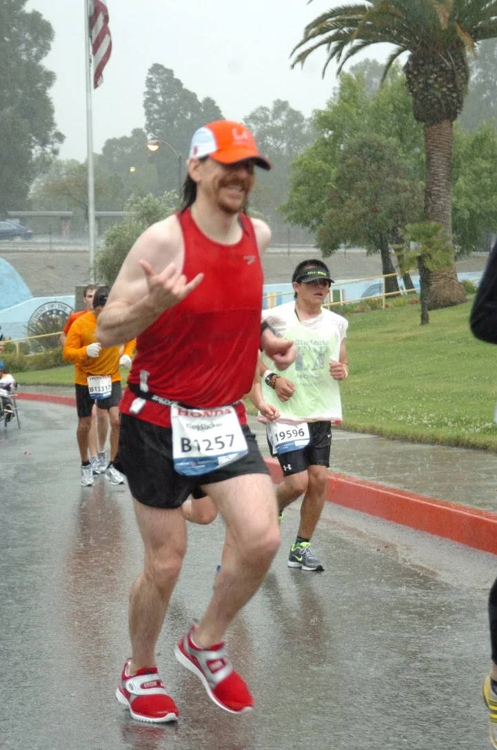 Runner in red tank top, black shorts, and red sneakers running in rain during a marathon, with others and trees in the background.