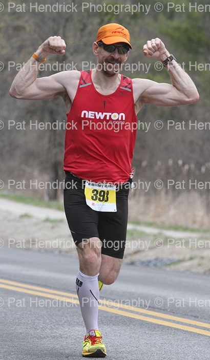Male marathon runner in red tank top, black shorts, orange cap, and sunglasses, flexing his biceps as he runs on a paved road outdoors.