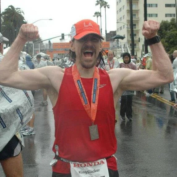 Man in a red sleeveless running shirt and a red cap, celebrating and flexing his biceps with a medal around his neck after a marathon in the rain.