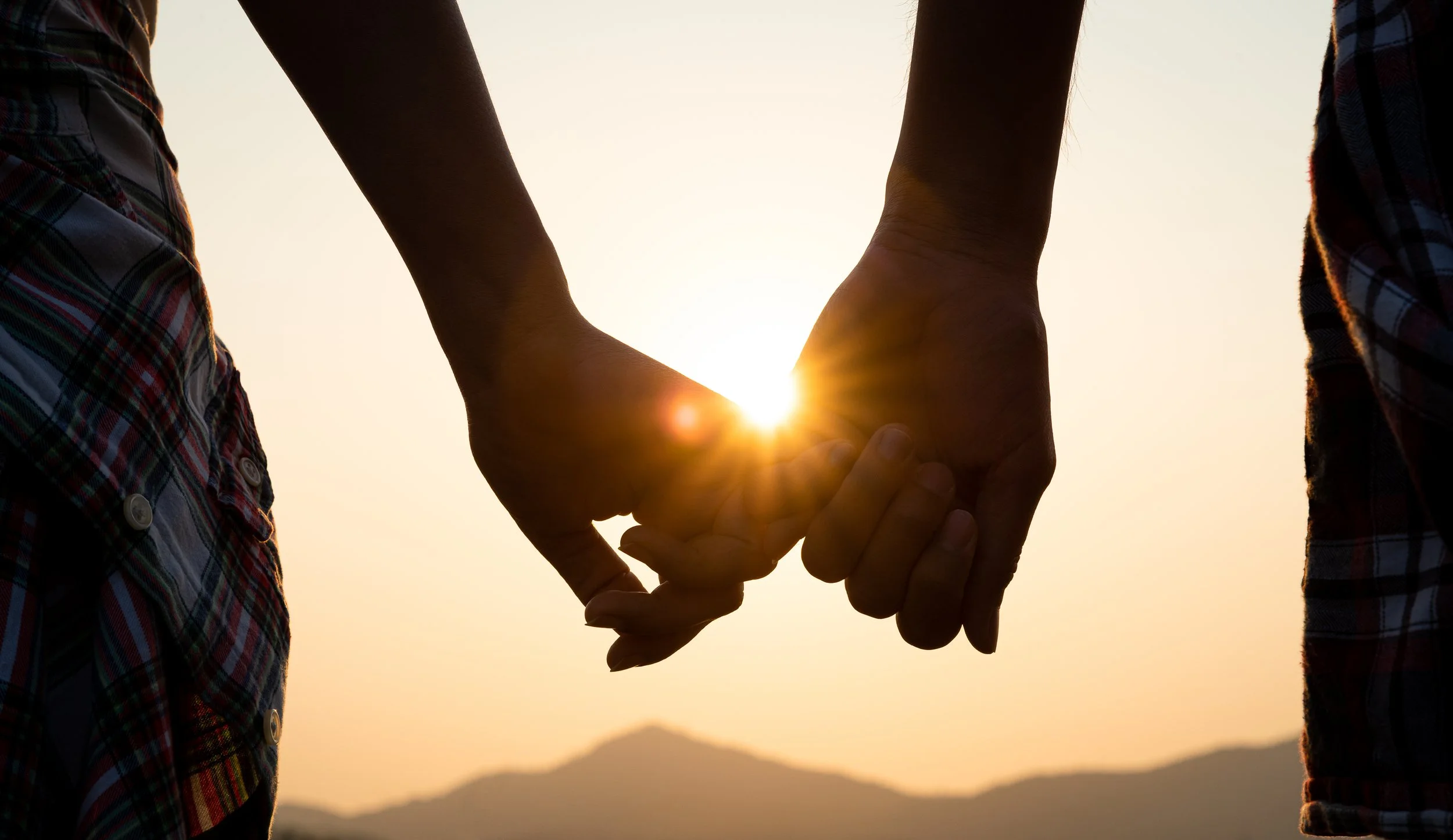 Two people holding hands with sunlight shining between their fingers, sunset or sunrise in the background, mountains in the distance.