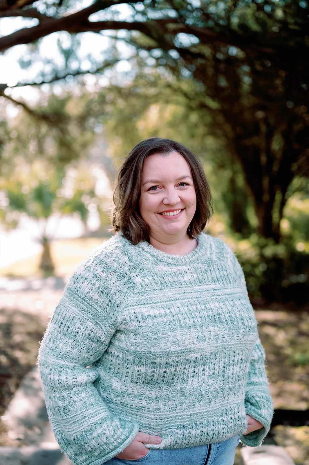 A woman with shoulder-length brown hair smiling outdoors in front of trees, wearing a light-colored knit sweater.