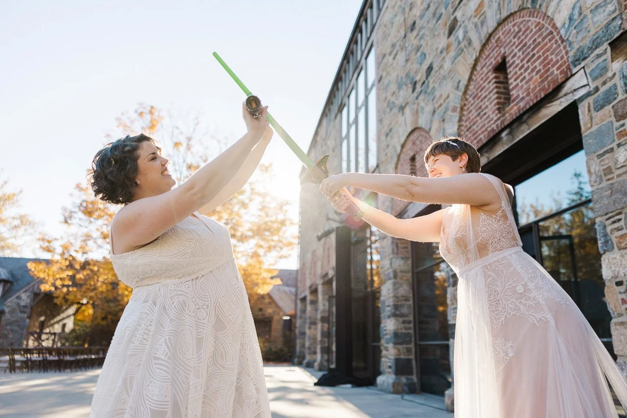 Two women in wedding dresses playing with a sword outside a stone building with large windows and trees in the background.
