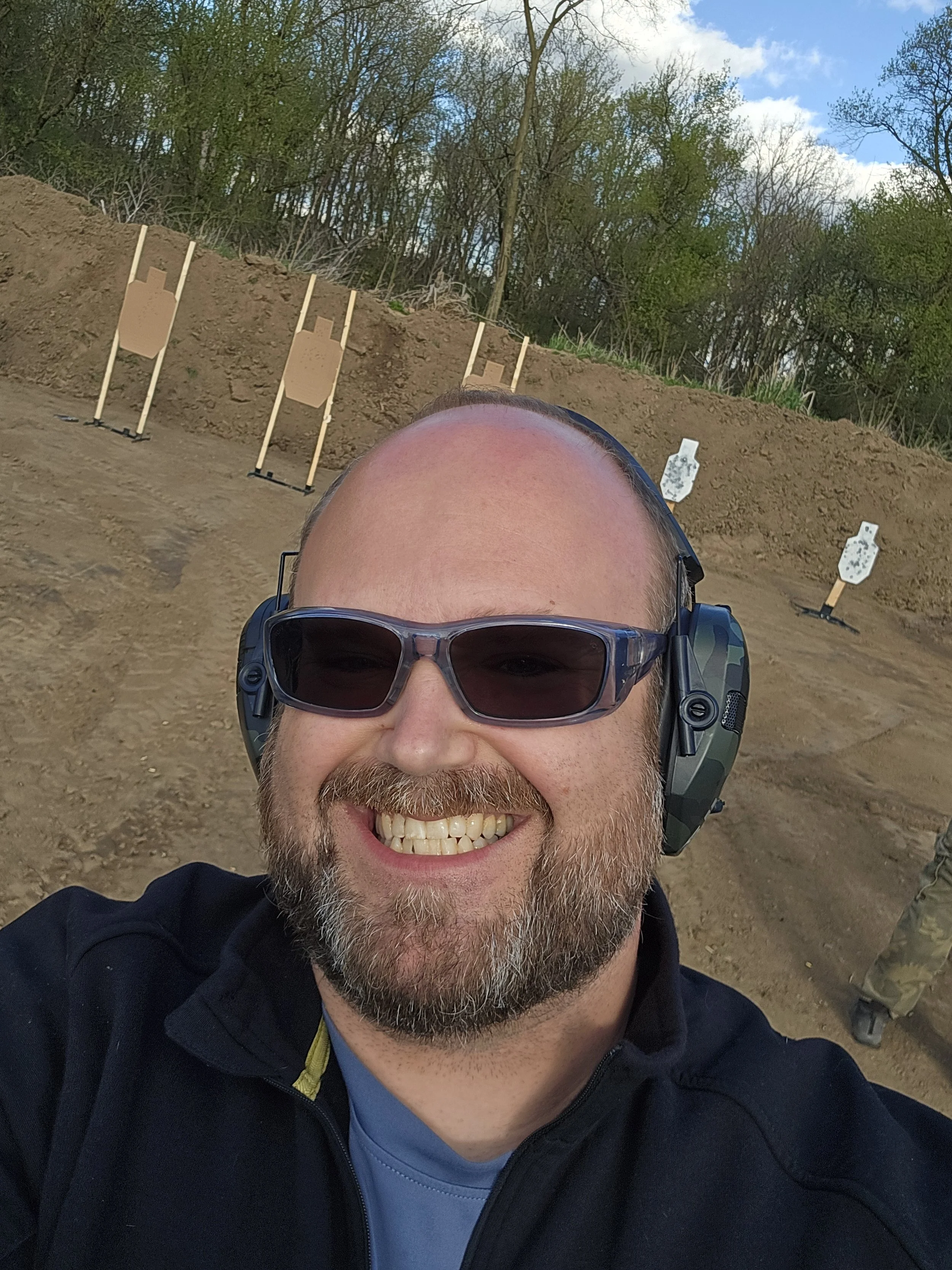 A man with a beard and sunglasses smiling at the camera, wearing a black jacket and a gray shirt, with a background of a dirt field, targets, and trees under a partly cloudy sky.