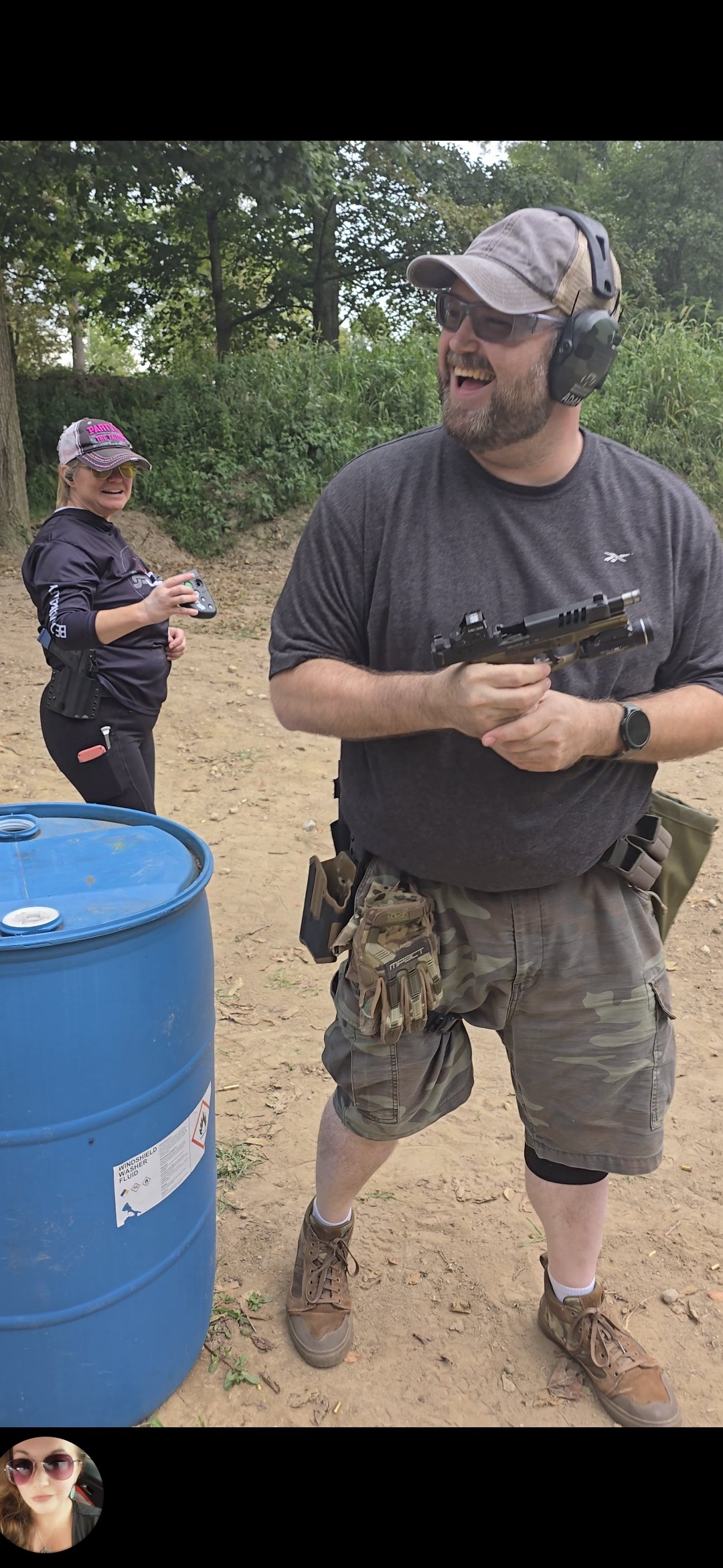 Man wearing sunglasses, cap, and camouflage shorts holding a handgun, smiling at an outdoor shooting range, woman in the background holding a handgun and wearing sunglasses and a cap, trees in the background, large blue barrel in the foreground.